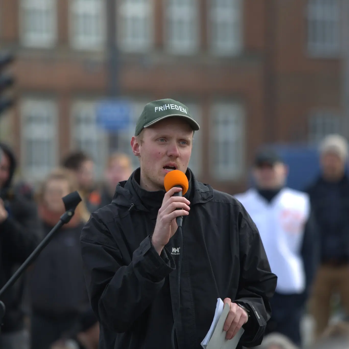 Billede af Lukas udenfor med orange mikrofon og kasket med teksten "Friheden"