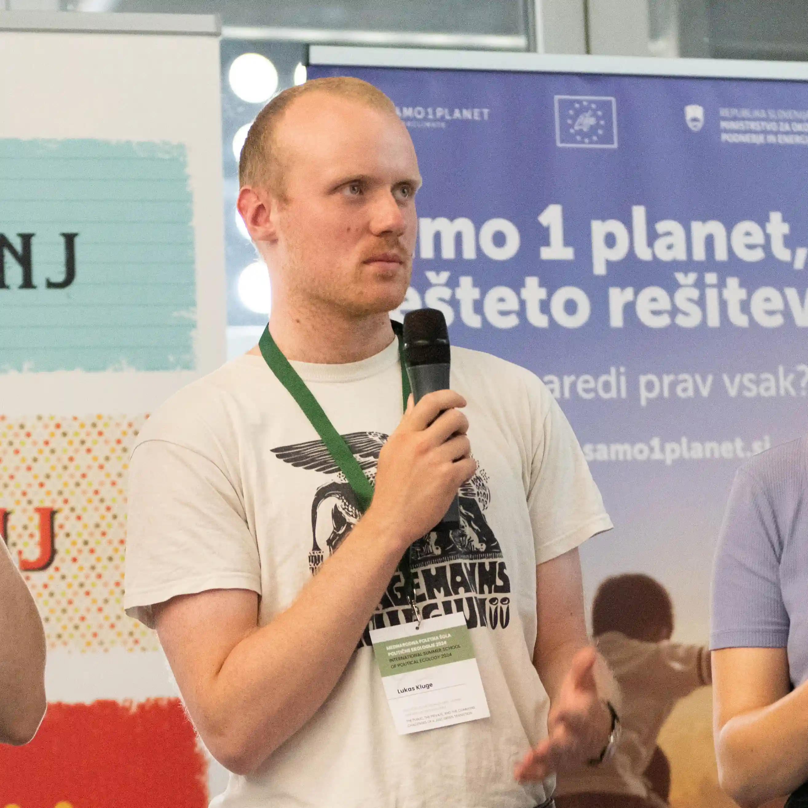 Billede af Lukas Kluge, en ung mand med kort hår, der står med en mikrofon. I baggrunden hænger der banner med tekst på slovensk